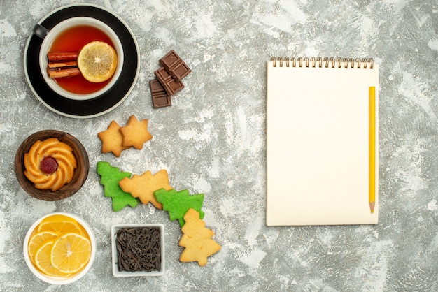 Vista superior de galletas de árbol de Navidad taza de cuencos de té con lápiz de cuaderno de rodajas de chocolate y limón sobre mesa gris