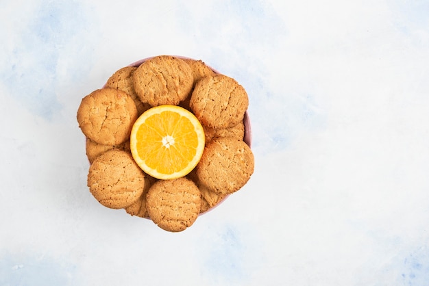 Vista superior de galletas caseras con media naranja cortada en un tazón sobre la mesa blanca.