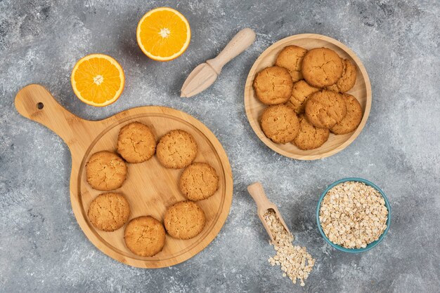 Vista superior de galletas caseras sobre tabla de madera y avena con naranjas sobre mesa gris.