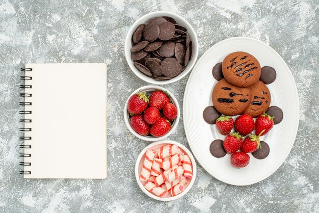 Vista superior de las galletas de chocolate, fresas y bombones redondos en el plato ovalado blanco y cuencos con dulces, fresas, chocolates y un cuaderno en el suelo blanco grisáceo