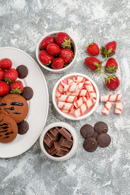 Vista superior de las galletas de fresas y bombones redondos en la placa ovalada blanca y tazones de dulces, fresas, chocolates en la mesa de color blanco grisáceo