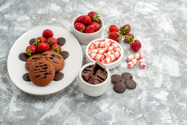 Foto gratuita vista superior de las galletas de fresas y bombones redondos en la placa ovalada blanca y tazones de dulces, fresas, chocolates en la mesa de color blanco grisáceo
