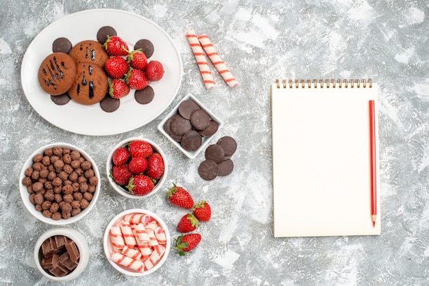 Vista superior galletas fresas y bombones redondos en la placa ovalada cuencos con dulces fresas chocolates cereales y un cuaderno con lápiz sobre la mesa gris-blanca