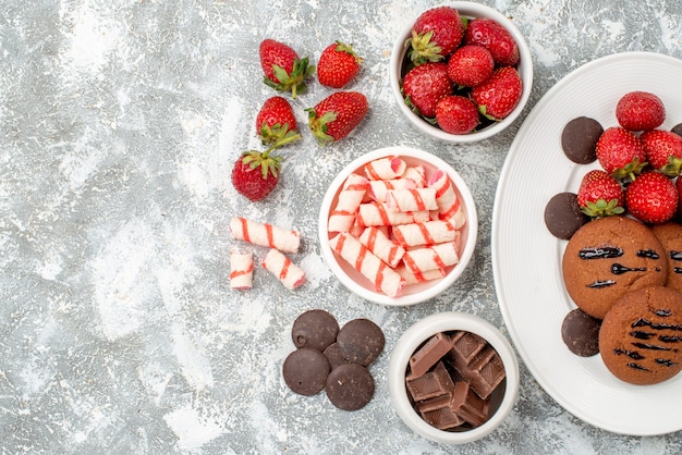 Vista superior galletas fresas y bombones redondos en la placa ovalada cuencos de dulces fresas chocolates en el lado derecho de la mesa gris-blanca