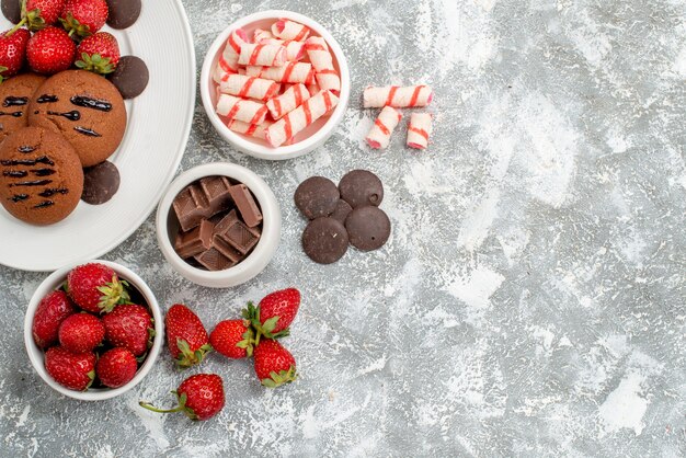 Vista superior de galletas, fresas y bombones redondos en el plato ovalado blanco, cuencos de dulces, fresas, chocolates en la parte superior izquierda de la mesa gris-blanca.