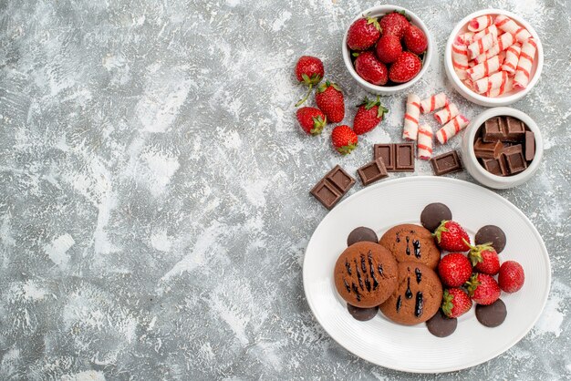 Foto gratuita vista superior de galletas de fresas y bombones redondos en el plato ovalado blanco y tazones de dulces, fresas, chocolates en el lado derecho de la mesa gris-blanca