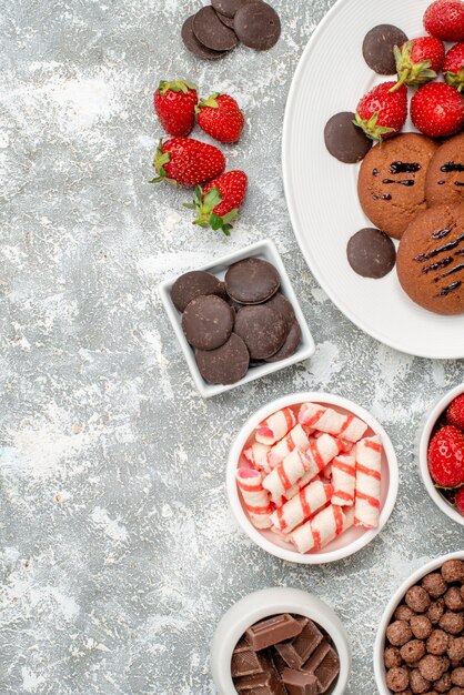 Vista superior galletas fresas y bombones redondos en el plato ovalado cuencos con dulces fresas chocolates cereales en el lado derecho de la mesa gris-blanca