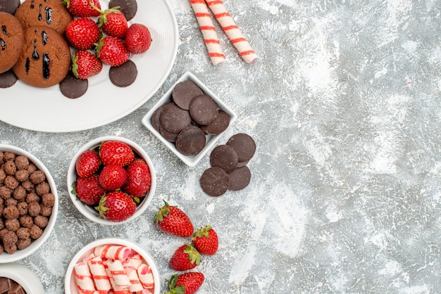 Foto gratuita vista superior galletas fresas y bombones redondos en el plato ovalado cuencos con dulces fresas chocolates cereales en el lado izquierdo de la mesa gris-blanca