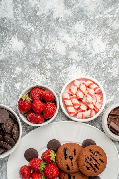 Foto gratuita vista superior de galletas, fresas y chocolates redondos en el plato ovalado blanco rodeado de cuencos con dulces, fresas y chocolates en la parte inferior de la mesa gris-blanca.