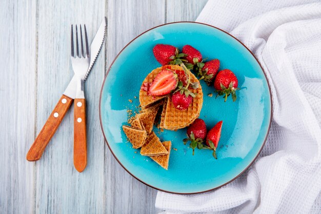 Vista superior de galletas de gofres y fresas en placa con tenedor y cuchillo sobre tela y superficie de madera