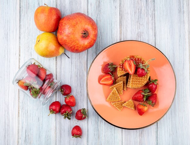 Vista superior de galletas de gofres y fresas en un plato con fresas derramando del tarro y granada de pera de manzana sobre superficie de madera