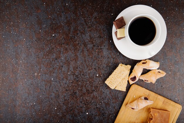 Vista superior de galletas de harina con mermelada de fresa en una tabla de madera y trozos de sésamo kozinaki y una taza de café sobre fondo negro con espacio de copia