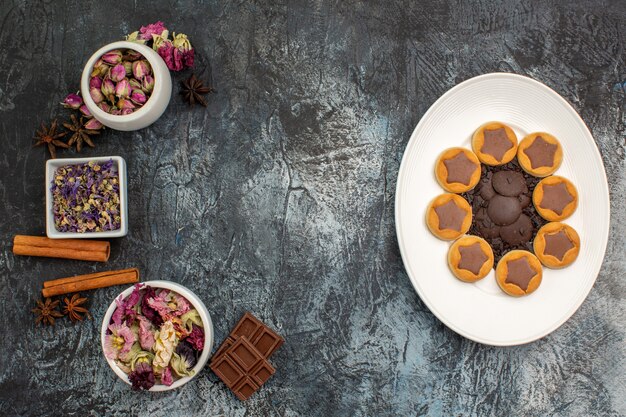 Vista superior de galletas en plato blanco y tazones de flores secas y chocolates sobre suelo gris