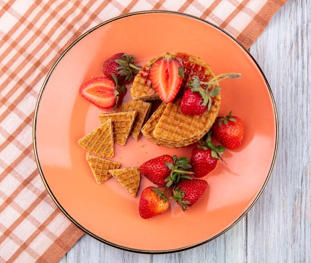 Vista superior de galletas waffle con fresas en placa sobre tela escocesa y superficie de madera