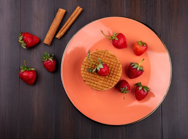 Vista superior de galletas waffle y fresas en un plato con canela sobre superficie de madera