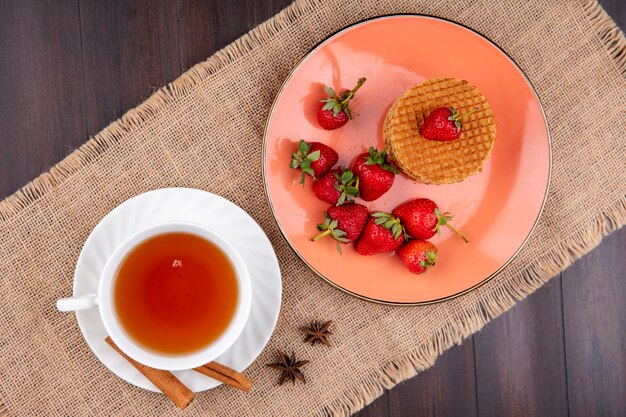 Vista superior de galletas waffle y fresas en plato y taza de té con canela en platillo sobre tela de saco y superficie de madera