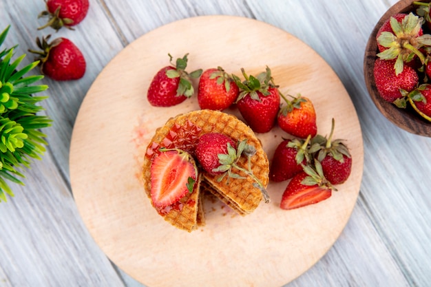 Vista superior de galletas waffle y fresas en plato y tazón y sobre superficie de madera