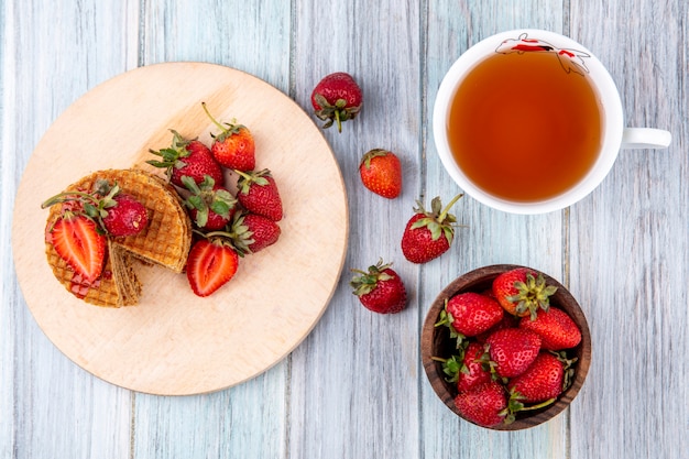 Vista superior de galletas waffle en tabla de cortar y fresas en un tazón con una taza de té en la superficie de madera