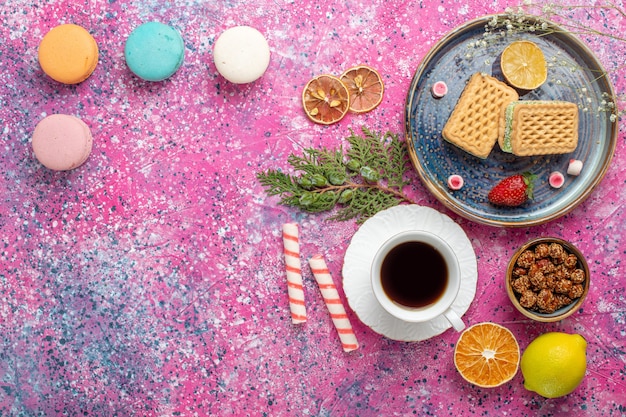 Vista superior de gofres dulces con taza de té y macarons en la superficie rosa