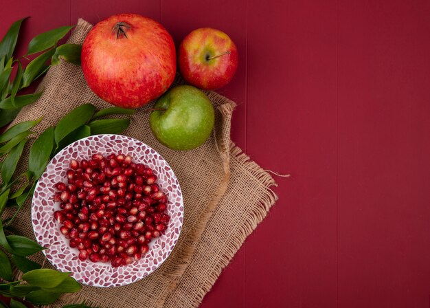 Vista superior de la granada pelada en un plato con manzanas y ramas de hojas sobre una superficie roja