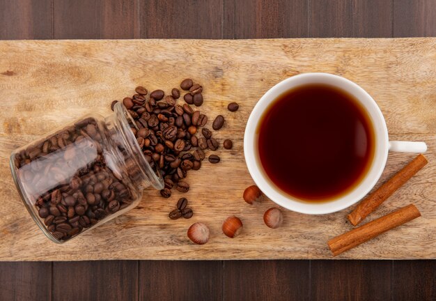 Vista superior de los granos de café que se derraman fuera del frasco de vidrio y una taza de té con canela y nueces en la tabla de cortar sobre fondo de madera