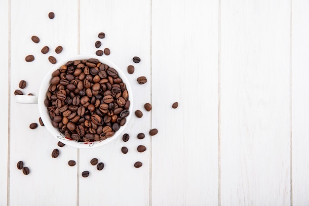 Vista superior de granos de café tostados oscuros en una taza blanca sobre un fondo de madera blanca con espacio de copia