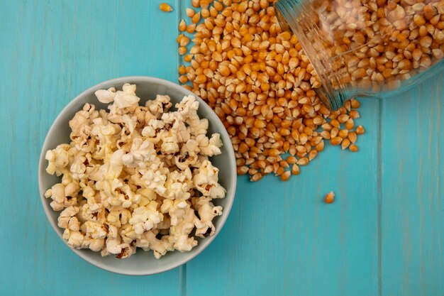 Vista superior de los granos de palomitas de maíz cayendo de un frasco de vidrio con sabrosas palomitas de maíz en un recipiente sobre una mesa de madera azul