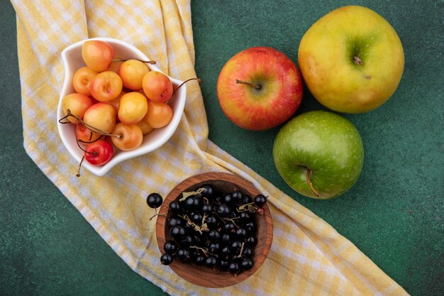 Foto gratuita vista superior de grosella negra con cerezas blancas en tazones sobre una toalla a cuadros amarilla con manzanas de colores sobre una superficie verde