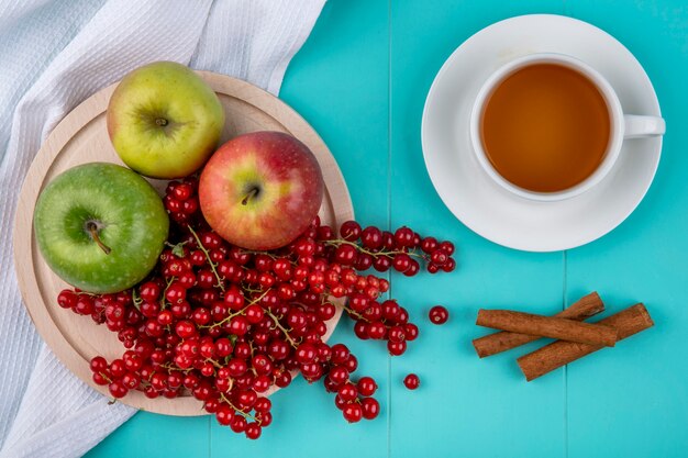 Vista superior de grosella roja con manzanas en un plato con canela y una taza de té sobre un fondo azul claro