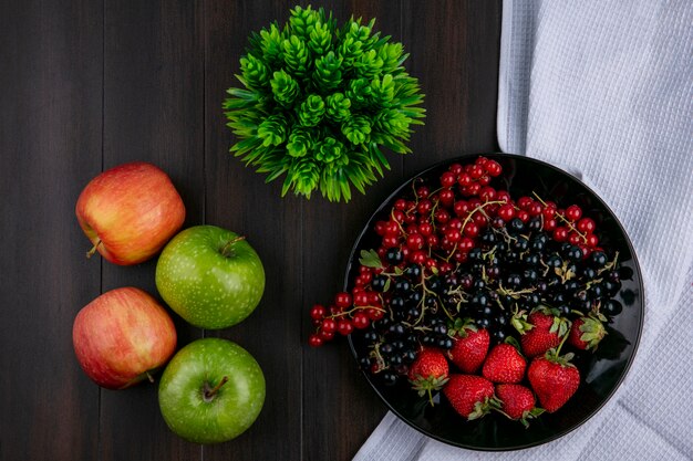 Vista superior grosellas rojas y negras con fresas en un plato con manzanas sobre un fondo de madera