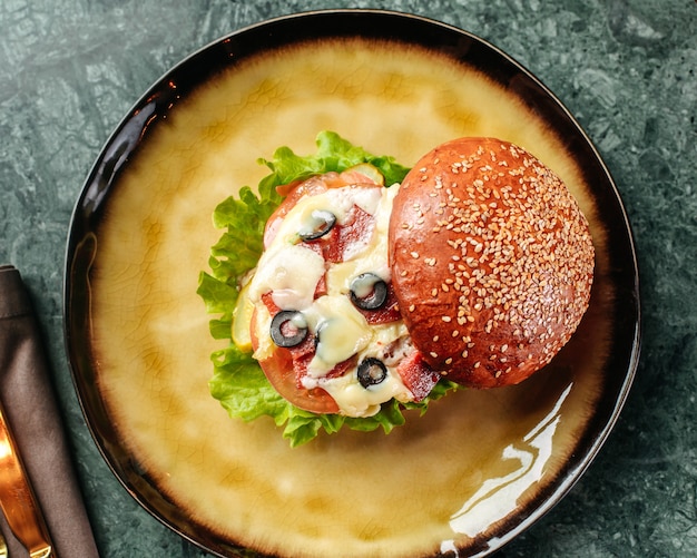 Una vista superior hamburguesa con aceitunas queso y diferentes verduras dentro de una sartén redonda en el piso brillante