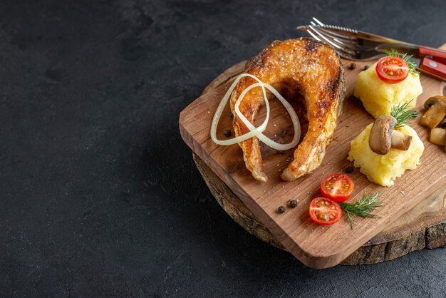 Vista superior de la harina de pescado frito con champiñones, verduras, queso y cubiertos en una tabla de madera en el lado izquierdo sobre una superficie angustiada negra