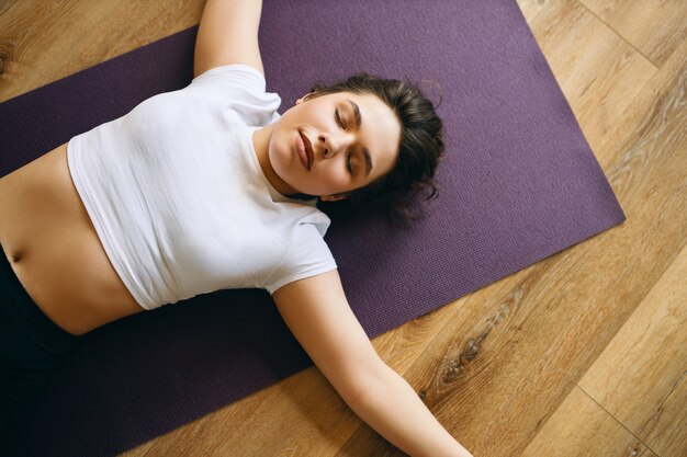 Vista superior de la hermosa mujer joven en top corto blanco en postura shavasana o cadáver durante la clase de yoga, descansando después de la práctica, meditando, respirando profundamente. Concepto de relajación y descanso