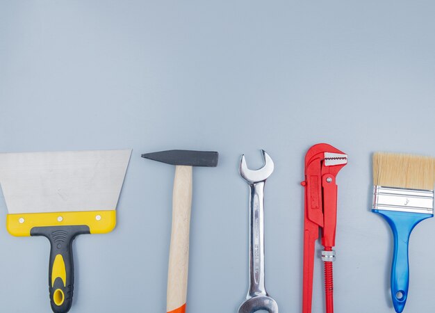 Vista superior de herramientas de construcción como martillo de ladrillo, llave de tubo, espátula, pincel y llave de boca abierta sobre fondo gris con espacio de copia