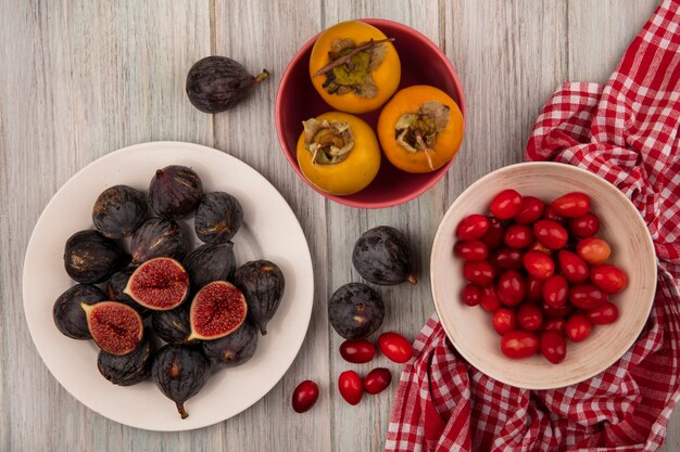 Vista superior de higos maduros de misión negra en un recipiente blanco con cerezas de cornalina en un recipiente sobre una tela marcada con frutos de caqui en una pared de madera gris