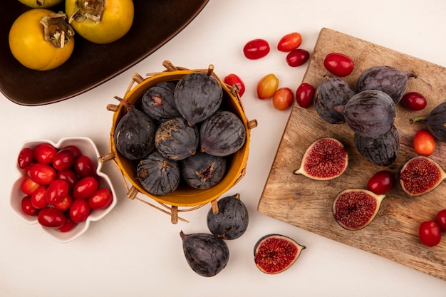 Vista superior de higos negros en un cubo con cerezas de cornalina en un recipiente con higos negros y cerezas de cornalina aislado en una tabla de cocina de madera en una pared blanca