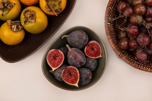 Vista superior de higos negros saludables en un recipiente con frutos de caqui en un recipiente sobre una pared blanca