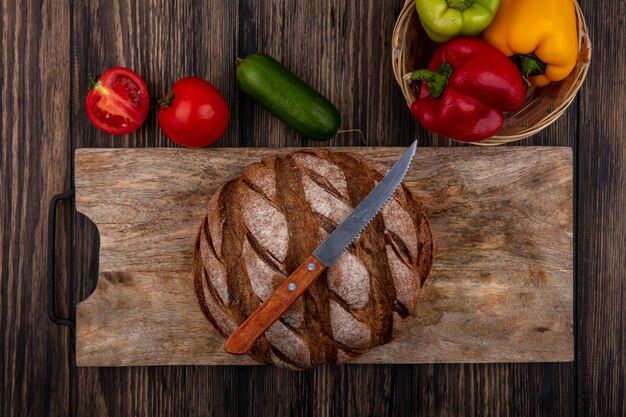 Vista superior de la hogaza de pan negro sobre un soporte con un cuchillo con tomates pepino y pimientos sobre un fondo de madera