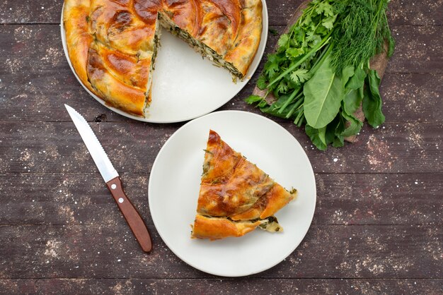 Vista superior de hojaldre de verduras cocidas en rodajas dentro de las placas con verduras frescas en el escritorio de madera marrón comida comida pastelería verde