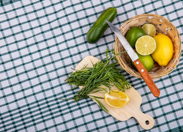 Vista superior de hojas de estragón sobre una tabla de cocina de madera con rodajas de limón con un cubo de limones frescos con un cuchillo sobre la superficie del mantel a cuadros