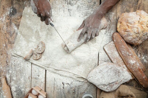 Vista superior del hombre afroamericano cocina cereales frescos, pan, salvado en la mesa de madera. Comida sabrosa, nutrición, producto artesanal.