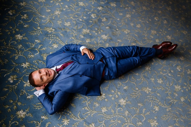 Vista superior del hombre feliz vestido con traje de moda y corbata roja sobre una alfombra azul con estampado de flores manteniendo la mano detrás de la cabeza posando y mirando a la cámara