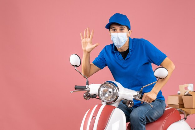 Vista superior del hombre de mensajería en máscara médica con sombrero sentado en scooter y mostrando cinco sobre fondo melocotón pastel