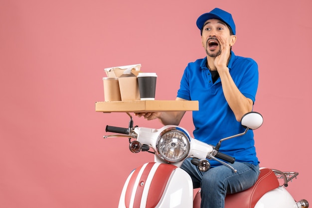 Vista superior del hombre de mensajería con sombrero sentado en scooter llamando a alguien sobre fondo melocotón pastel