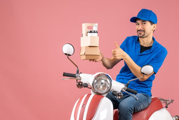Vista superior del hombre de mensajería con sombrero sentado en scooter mostrando pedidos haciendo gesto ok sobre fondo de melocotón pastel