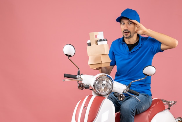 Vista superior del hombre de mensajería con sombrero sentado en scooter mostrando pedidos sintiéndose confundidos sobre fondo melocotón pastel