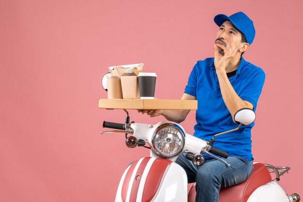 Vista superior del hombre de mensajería con sombrero sentado en scooter pensando profundamente sobre fondo melocotón pastel