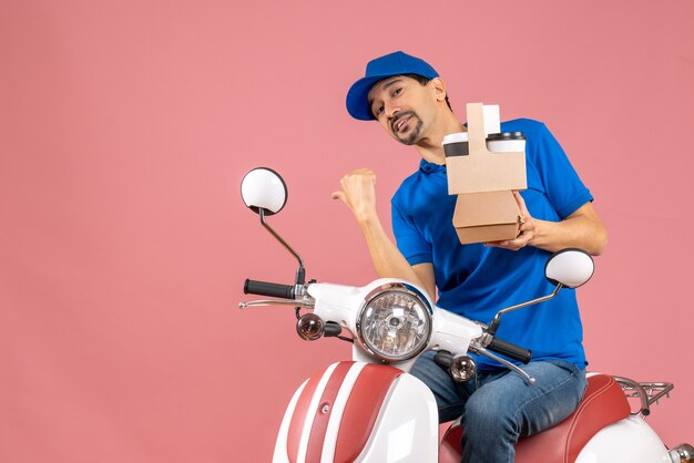 Vista superior del hombre de mensajería con sombrero sentado en scooter sosteniendo pedidos apuntando hacia atrás sobre fondo de melocotón pastel