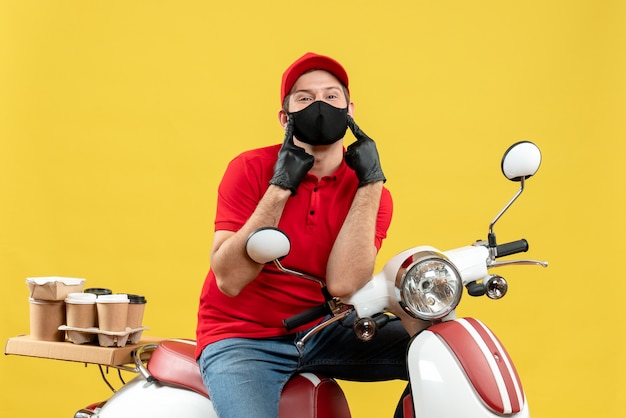 Vista superior del hombre de mensajería vestido con blusa roja y guantes de sombrero en máscara médica entregando orden sentado en scooter apuntando a sí mismo