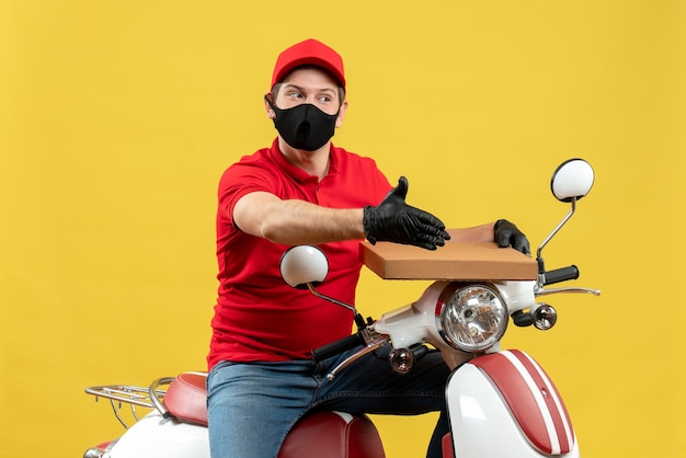 Vista superior del hombre de mensajería vestido con blusa roja y guantes de sombrero en máscara médica sentado en scooter mostrando orden dando la bienvenida a alguien